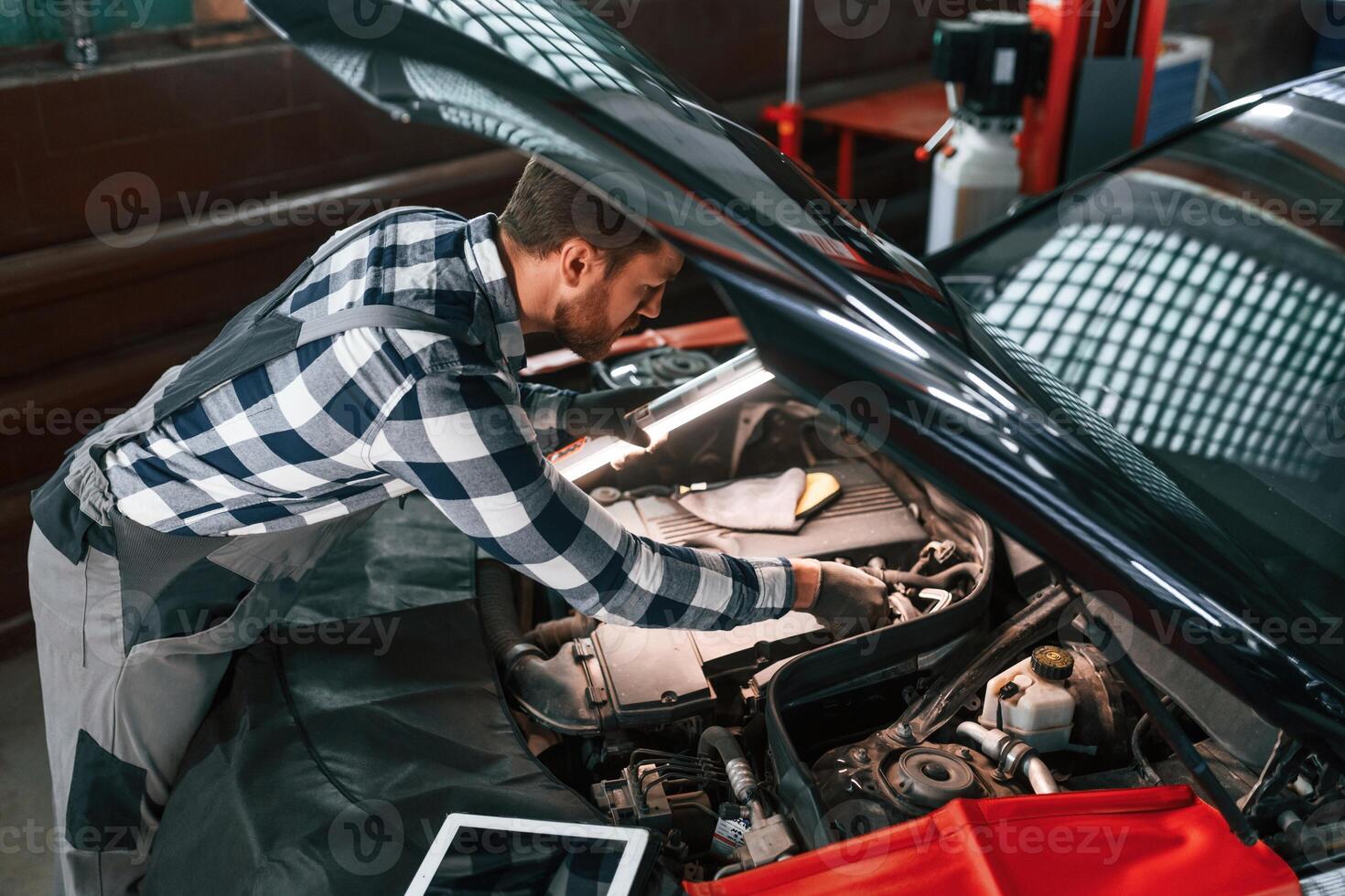 Under the hood.. Holding lighting equipment and fixing the issues of the car. Man in uniform is working in the auto salon photo