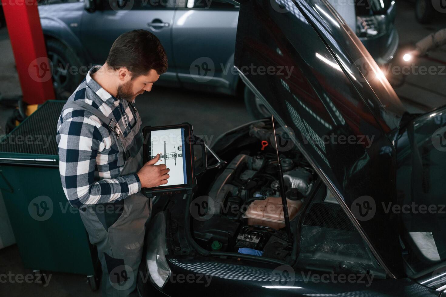 Standing near the car with hood opened. Holding tablet. Man in uniform is working in the auto salon photo