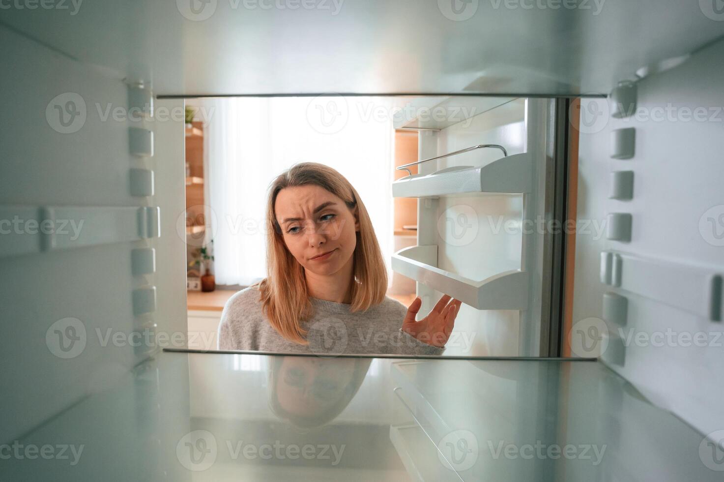 Disappointed about an empty fridge with no .food in it. Beautiful young woman is on the kitchen photo