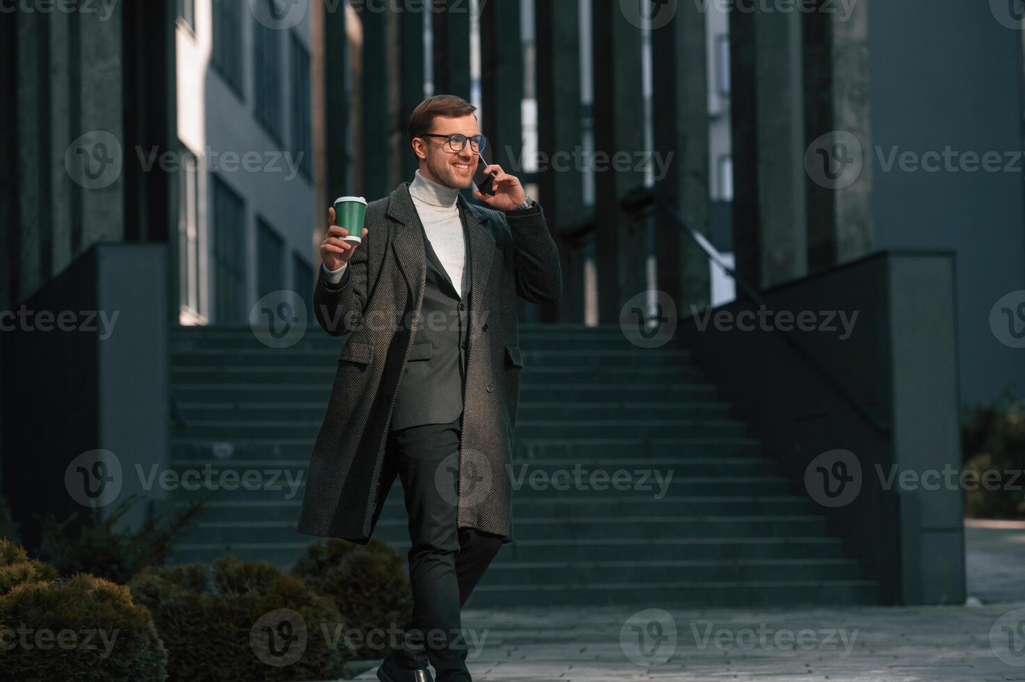 Amazing sunlight, holding, smartphone and cup of drink. Handsome man in formal clothes is outdoor near the business building photo