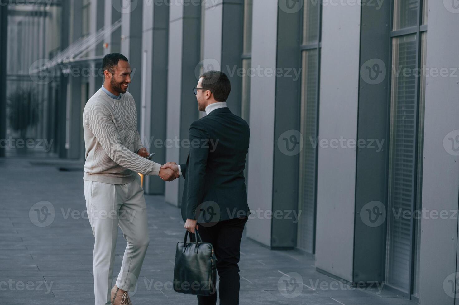 Doing, handshake, greetings gesture. Businessman in formal clothes is with black man outdoors in the city photo