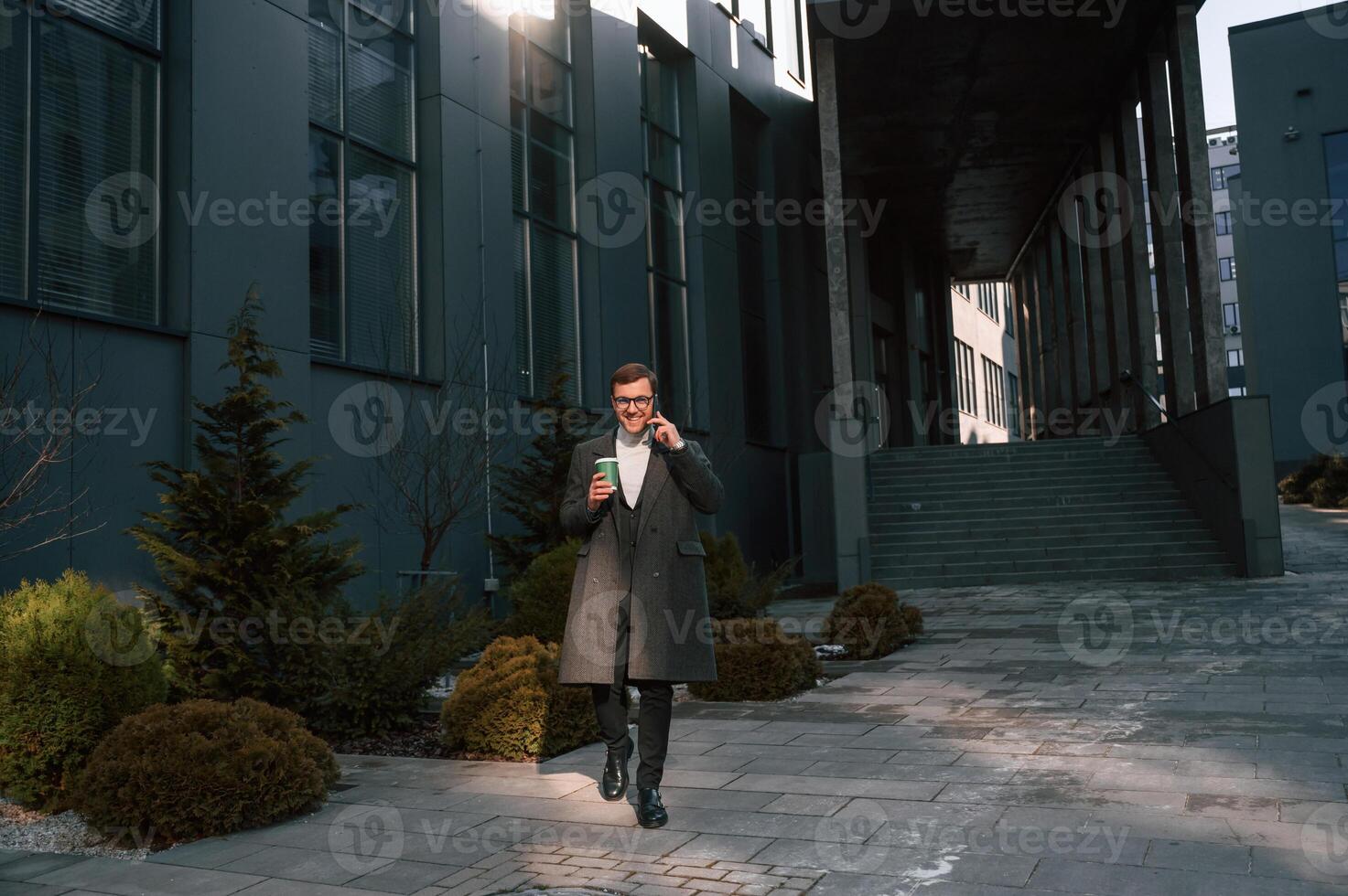 With cup of drink and smartphone. Handsome ,man in formal clothes is outdoor near the business building photo