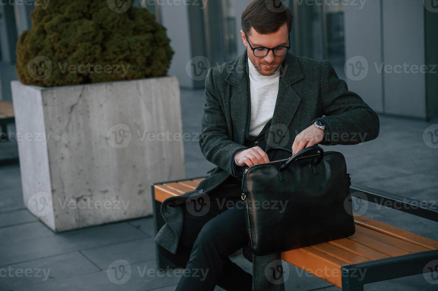 At daytime. On the bench. Handsome man in formal clothes is outdoor near the business building photo