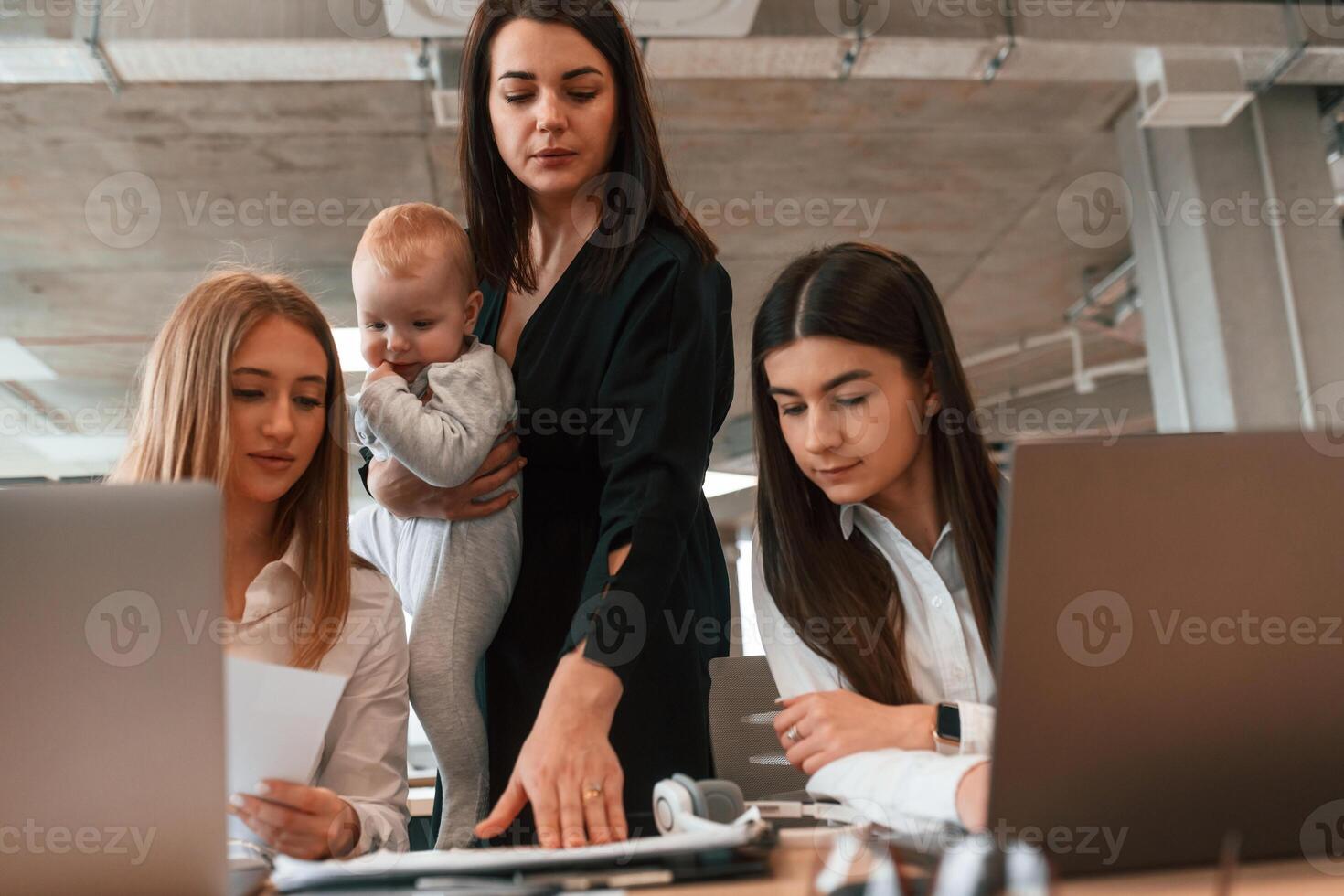Woman is standing with son in hands.. Infant baby is in the office where group of people are working together photo