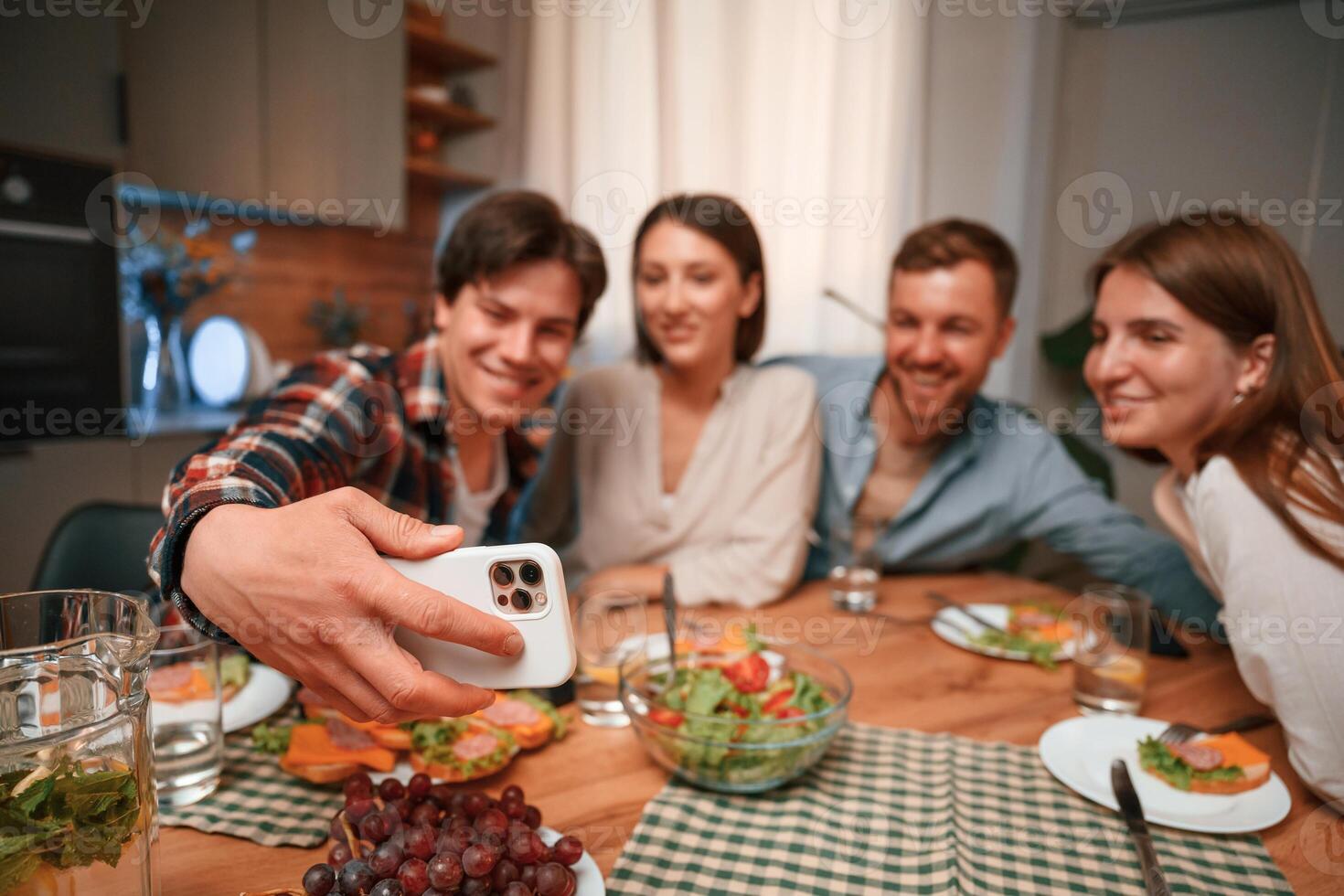 Man is making selfie. Group of people are on the kitchen, having dinner, spending time together photo
