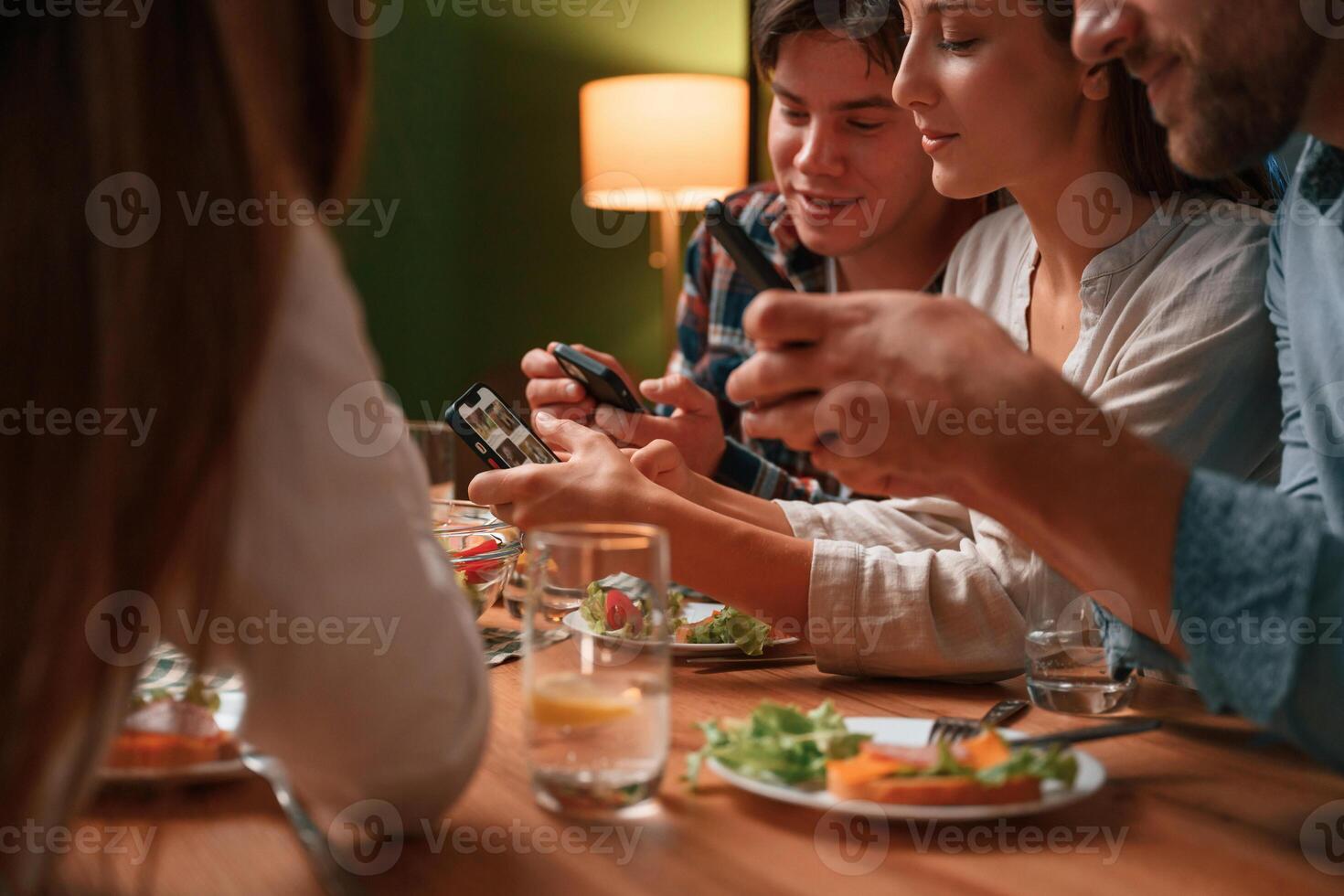 Sitting, by the table with smartphones in hands. Group of people are on the kitchen, having dinner, spending time together photo