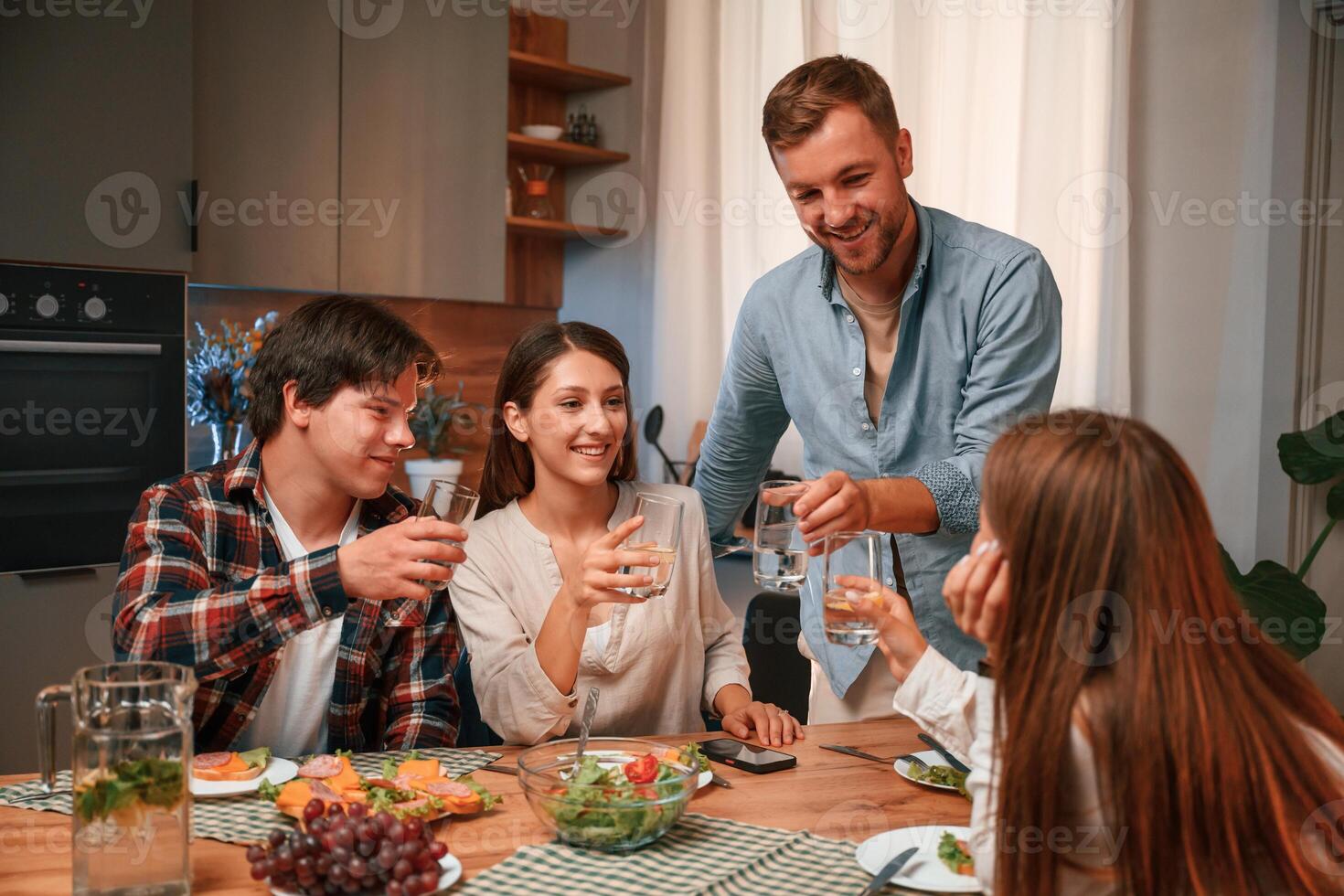 Holding, glasses with water. Group of people are on the kitchen, having dinner, spending time together photo