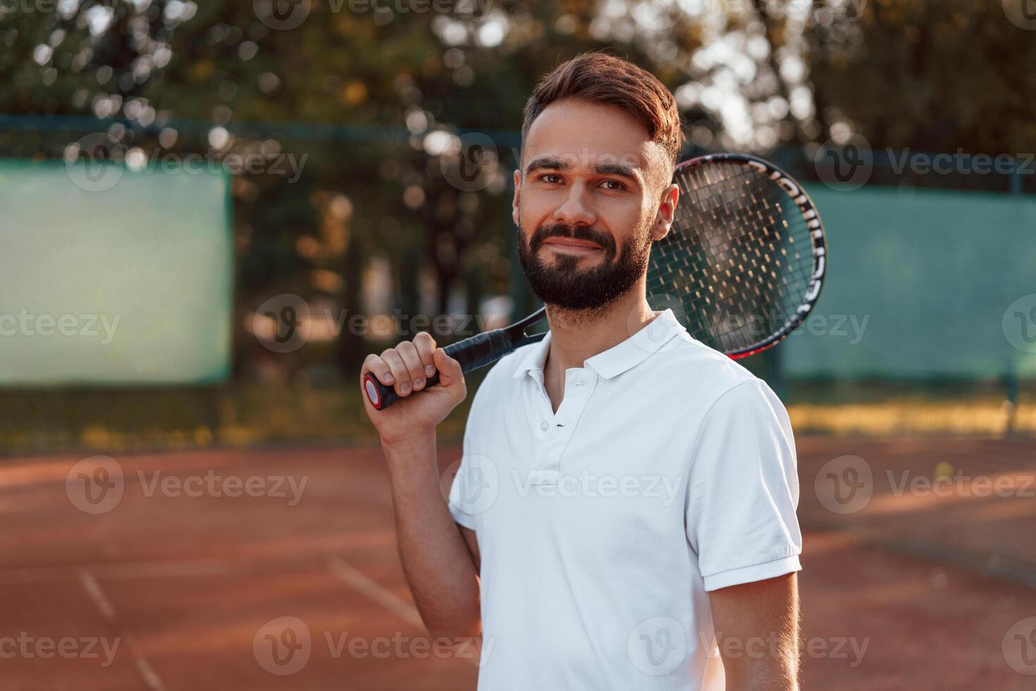 Positive, young, man is on the tennis court at sunny daytime. holding racket photo