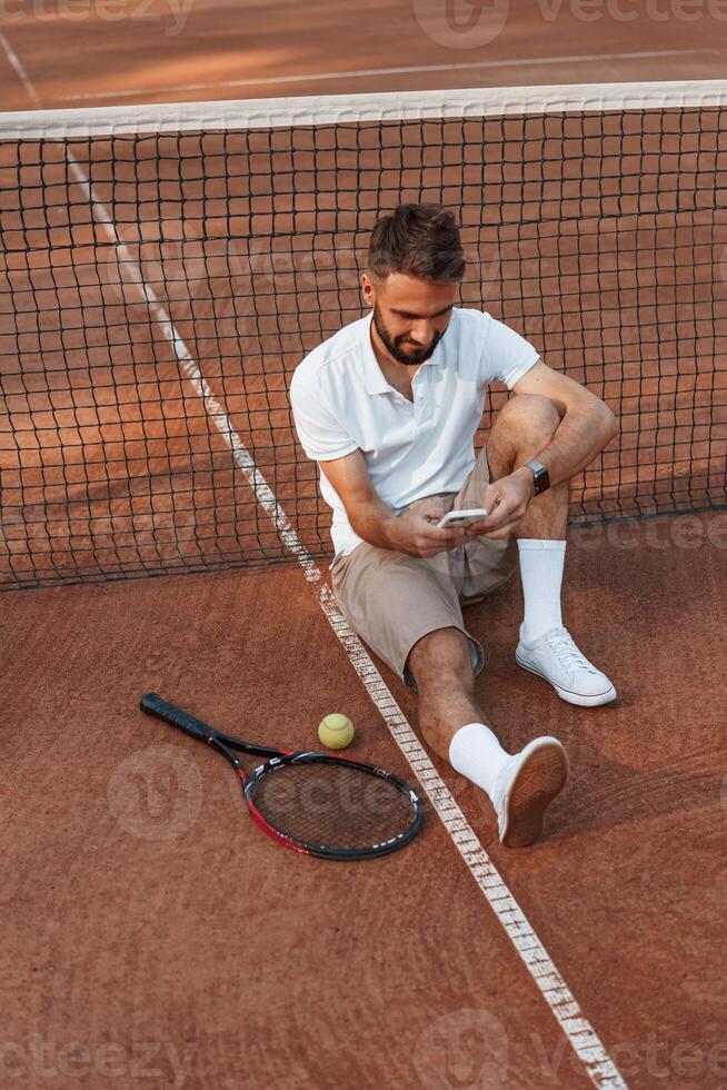 Taking a break. Sitting near the net. Young, man is on ,the tennis court at sunny daytime photo