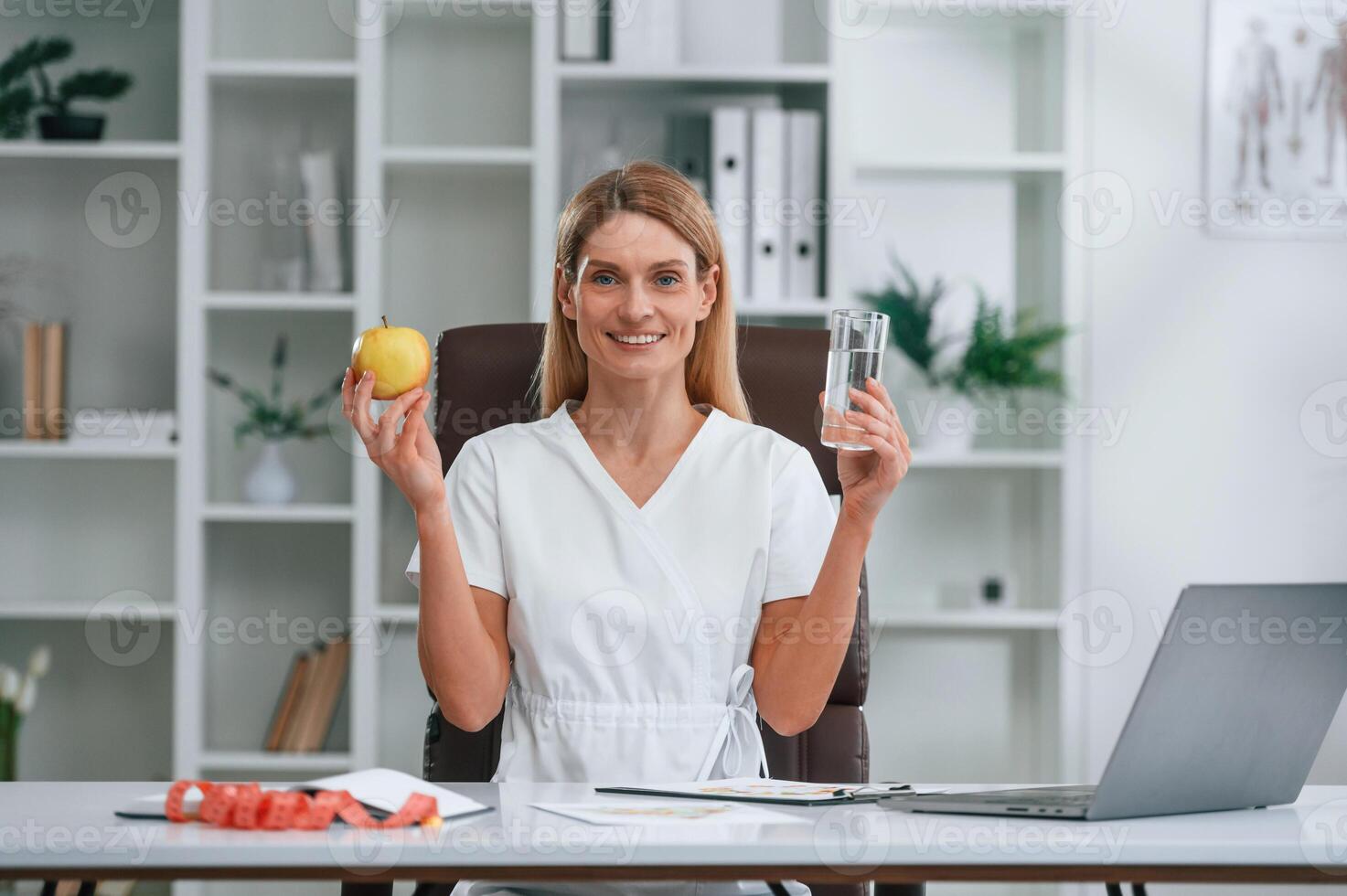 Healthy, eating, holding an apple and glass of water. Conception of diet. Young female doctor in white coat is indoors photo