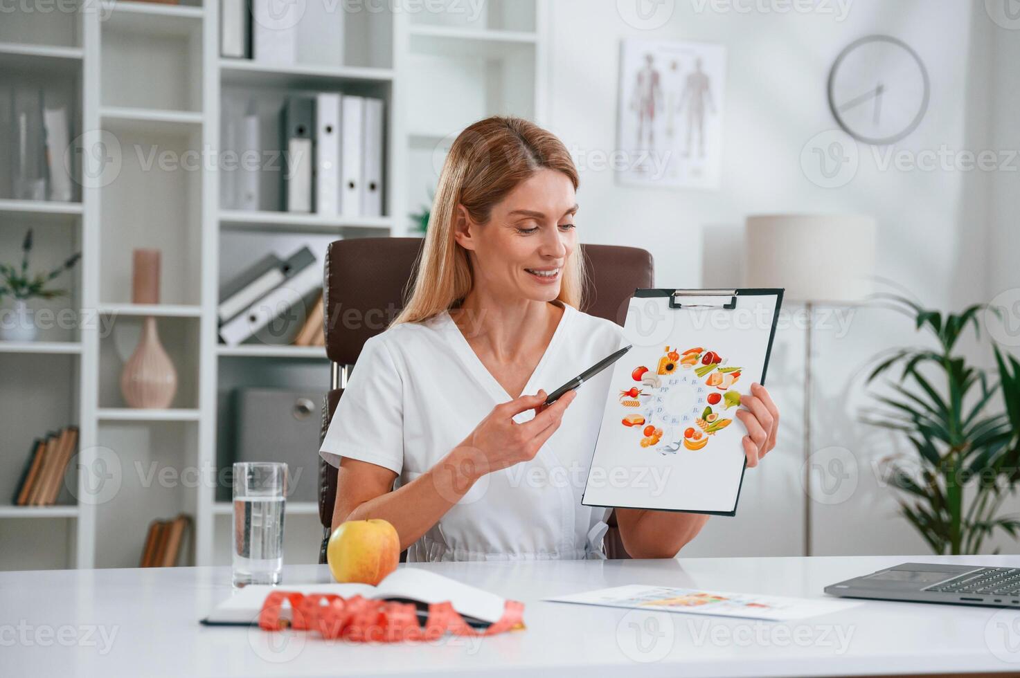 With picture of food and vitamins,. Young female doctor in white coat is indoors photo