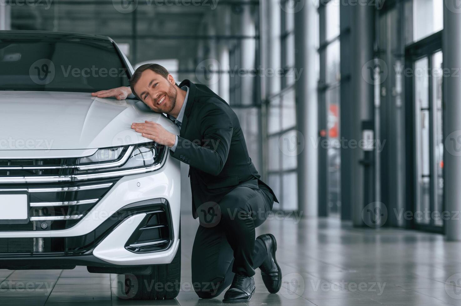 Embracing the ,vehicle. Man in black suit is indoors in the car dealership near automobile photo