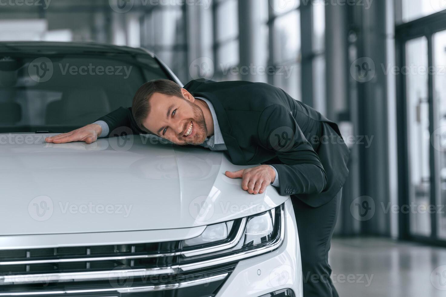 Embracing, the vehicle. Man in black suit is indoors in the car dealership near automobile photo