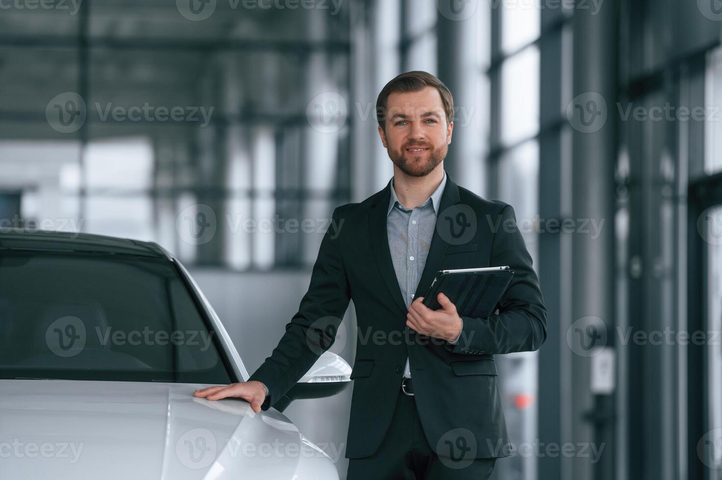 With, tablet in hands. Man in black suit is indoors in the car dealership near automobile photo