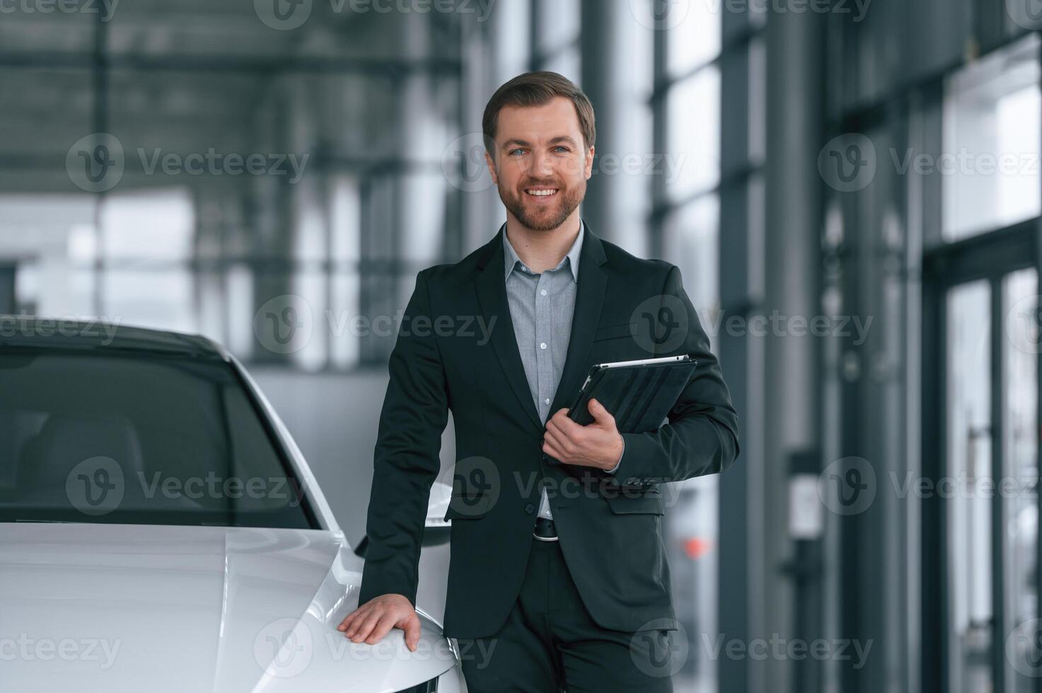 With tablet in hands.. Man in black suit is indoors in the car dealership near automobile photo