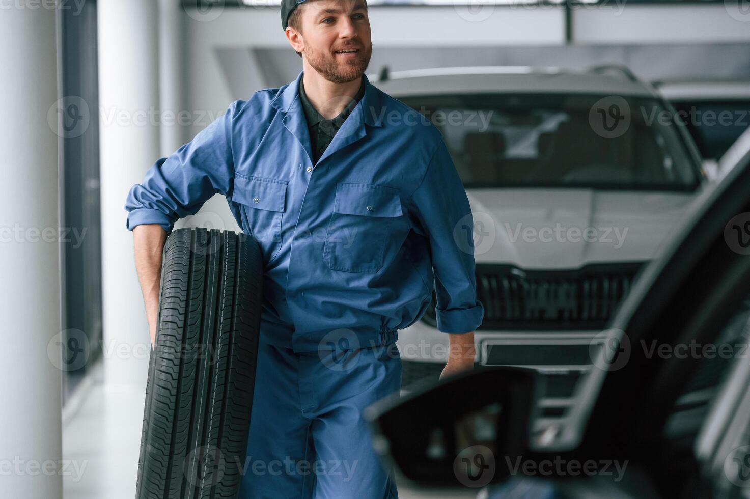Walking, forward ,and holding the tire. Man in blue uniform is working in the car service photo