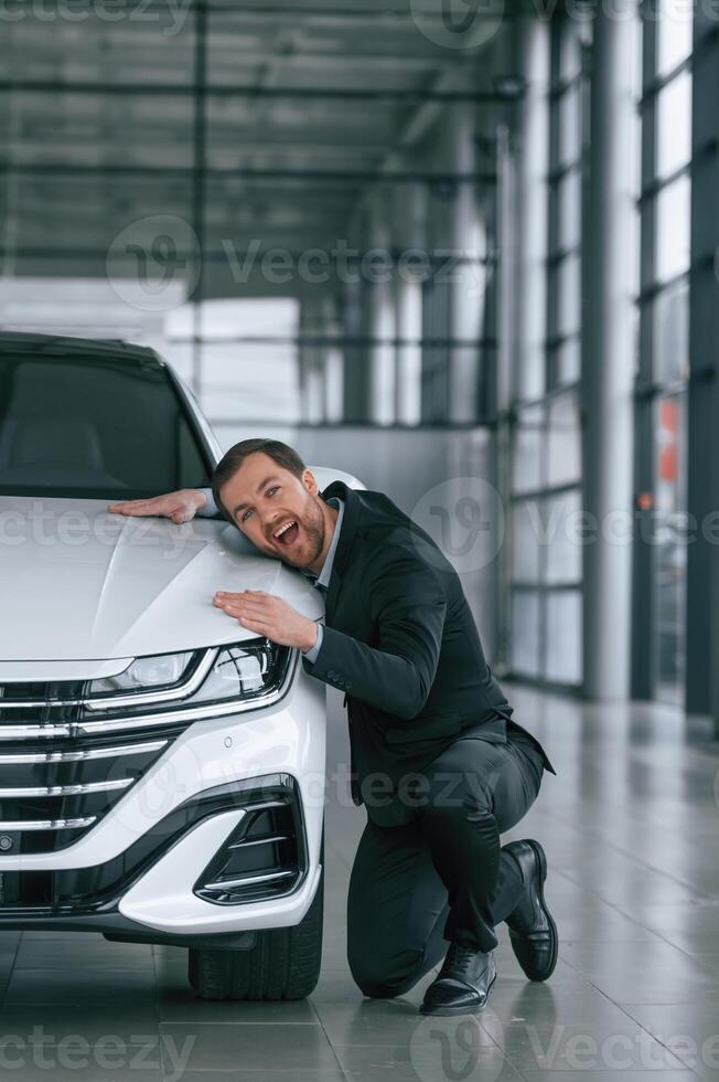 Embracing the vehicle. Man in black suit is indoors in the car dealership near automobile photo
