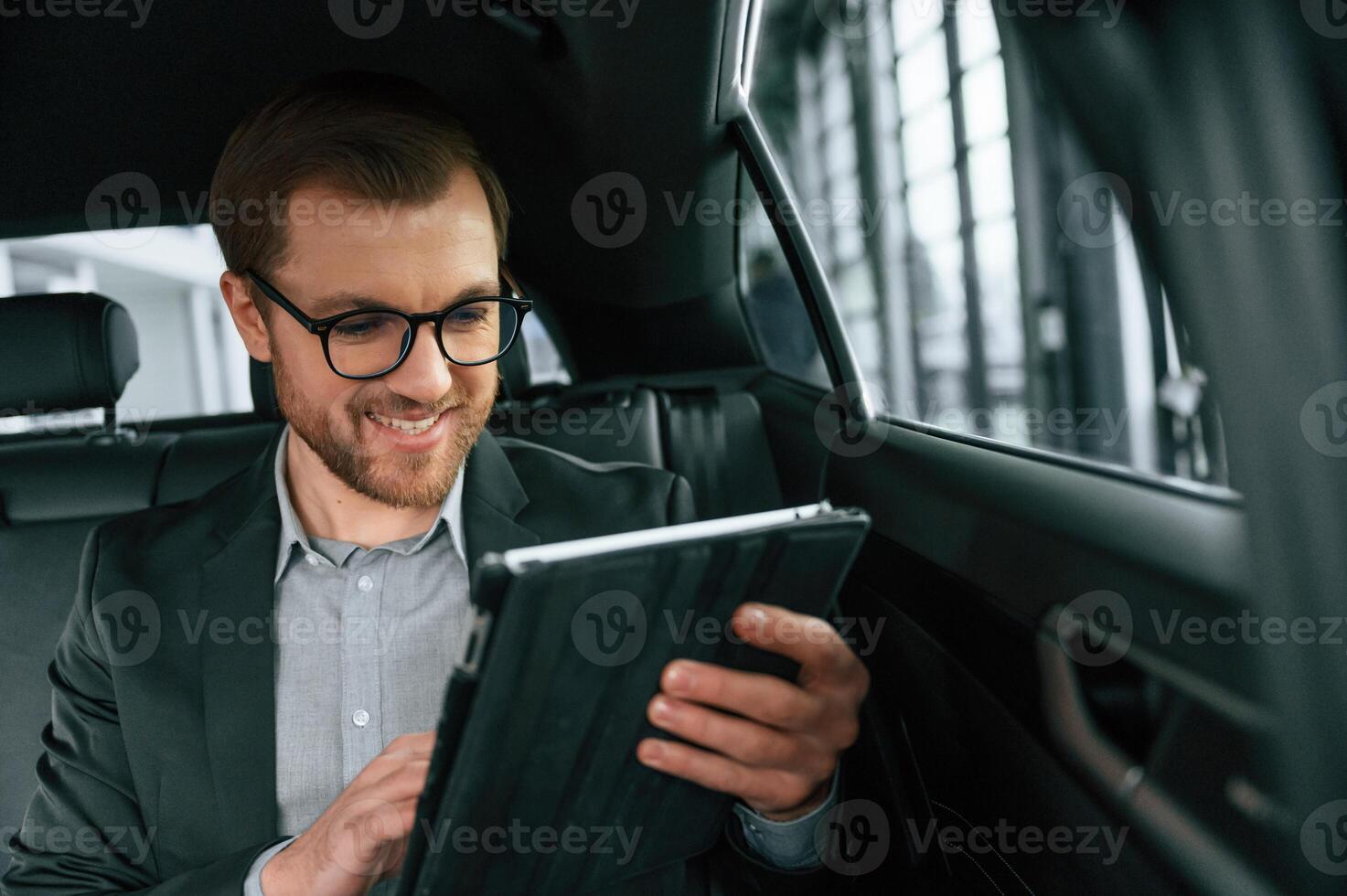 Smiling, using, tablet. Man in formal business clothes is sitting in the modern automobile photo