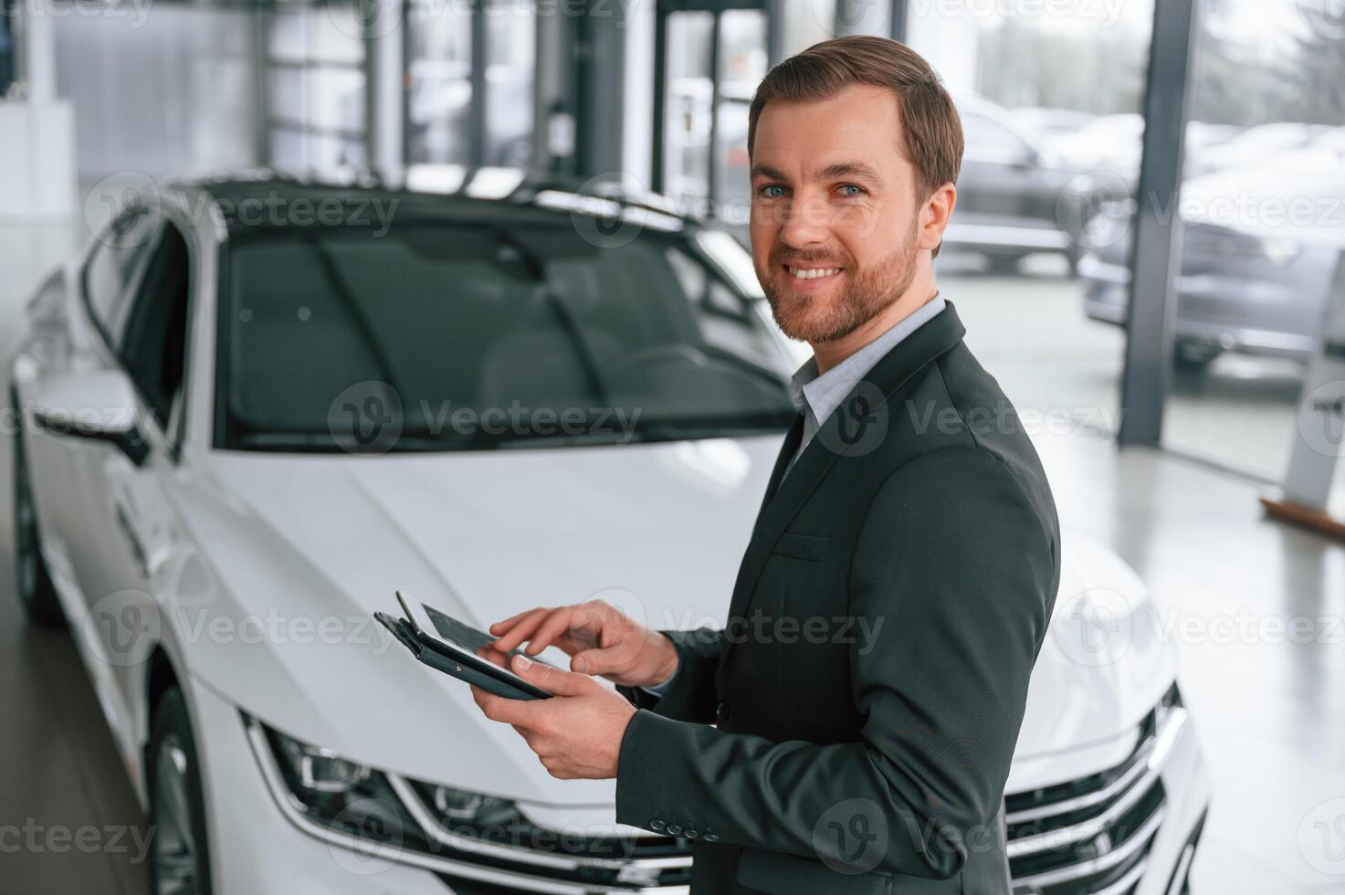 Standing,, side view, holding tablet. Man in, black suit is indoors in the car dealership near automobile photo