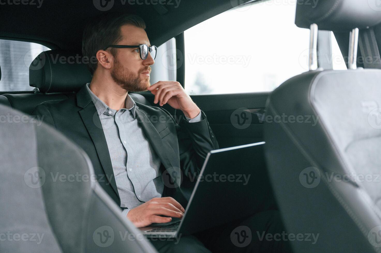 In glasses,, working by using laptop.. Man in formal business clothes is sitting in the modern automobile photo