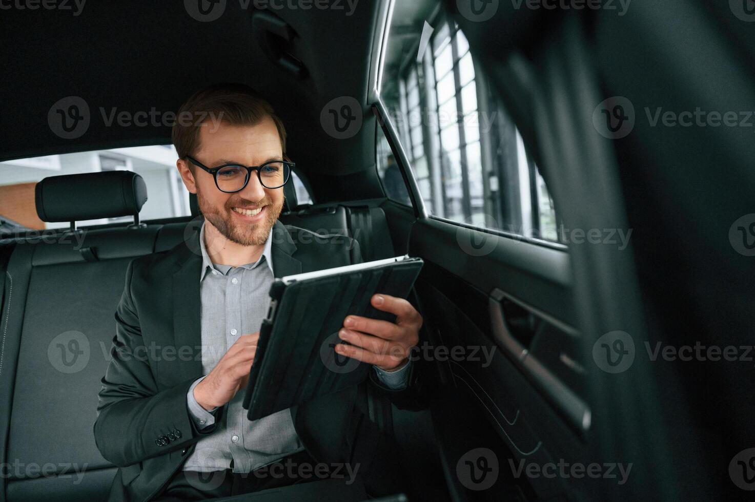 Smiling, using tablet. Man, in formal business, clothes is sitting in the modern automobile photo