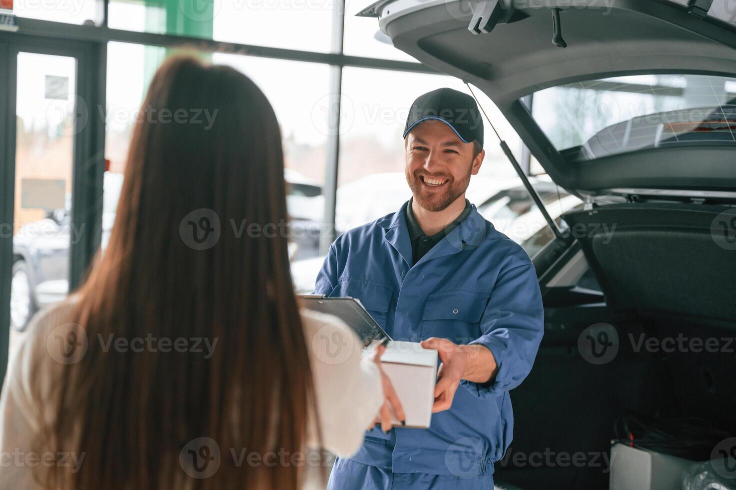 Man is, holding box and notepad. Young woman is visiting auto mechanic in garage. Repair service photo