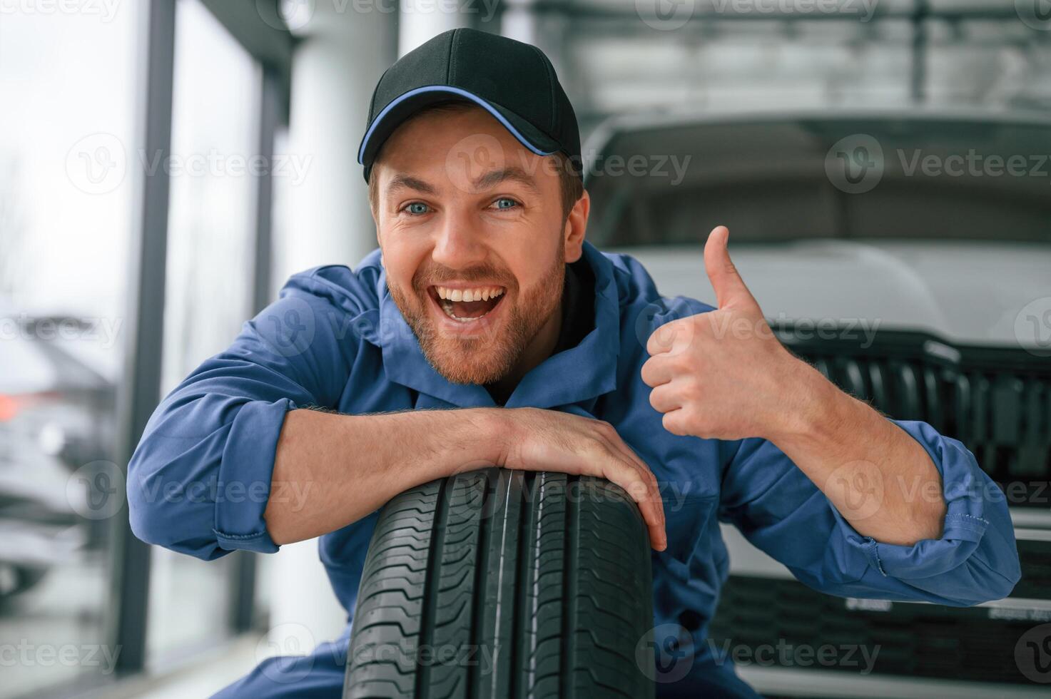 Leaning on the, tire and, smiling. Man in blue uniform is working in the car service photo