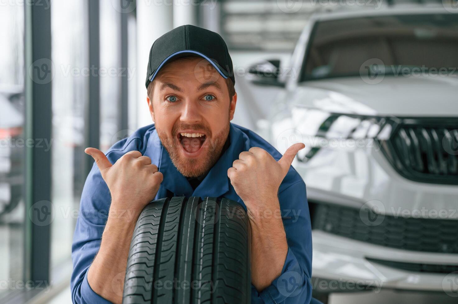 Leaning on the tire and smiling. Man in blue, uniform is working in the car service photo