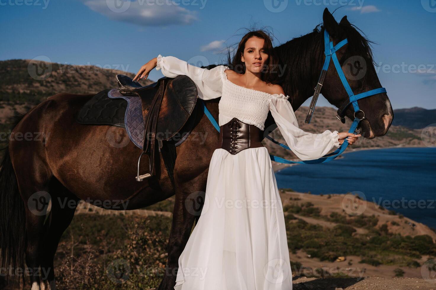 A young girl rider poses next to her horse against the backdrop of a mountain and sea landscape. The concept of riding. Artistic photography. Ready-made cover for books and magazines. photo