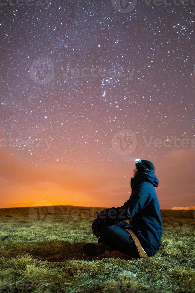 Side view of a man sitting on the grass wearing a jacket and hat, looking up at the milky way, using a headlamp photo
