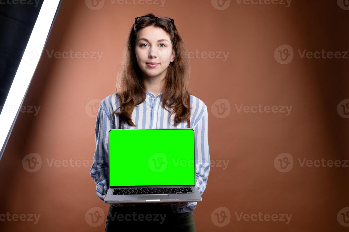 Caucasian cute girl presenting laptop with green blank screen for mockup, on brown studio wall background, dressed in office clothes photo