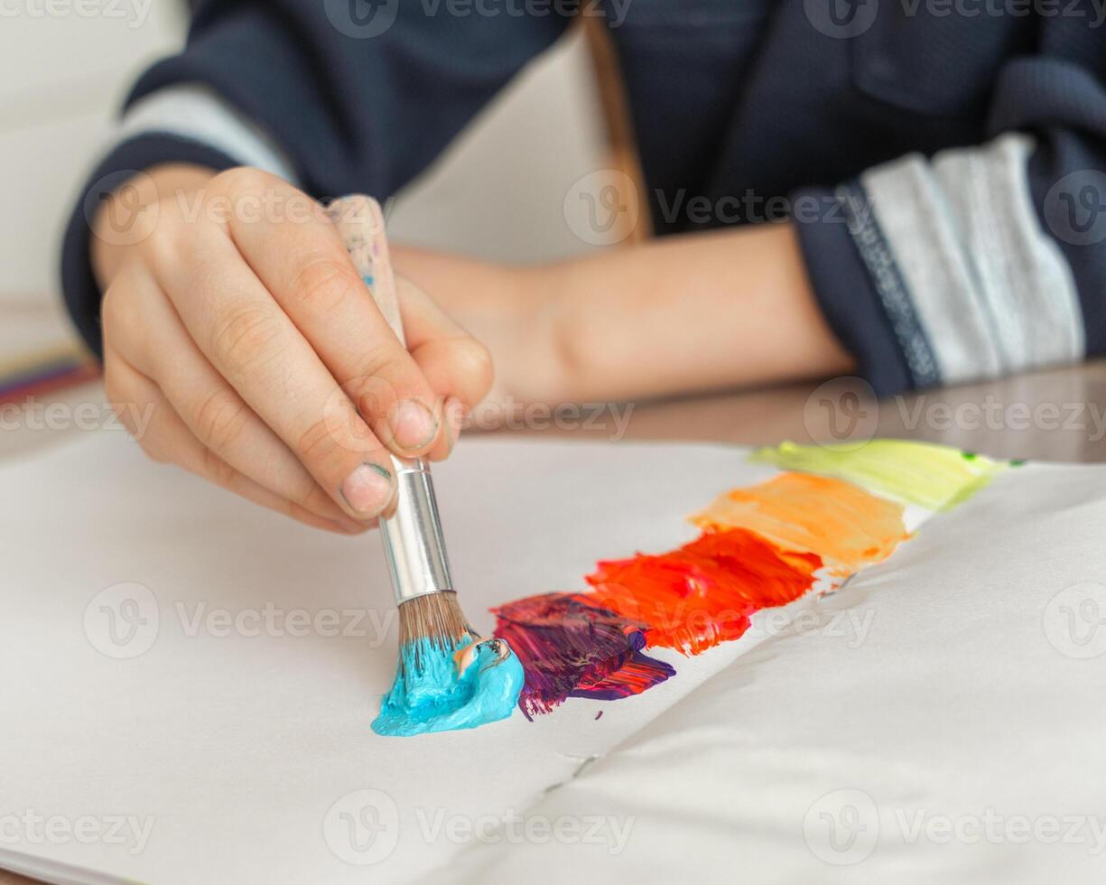 Boy's hand holds a brush with blue paint and draws a rainbow on a sheet of paper, a boy of 4-5 years old draws with paints at home photo