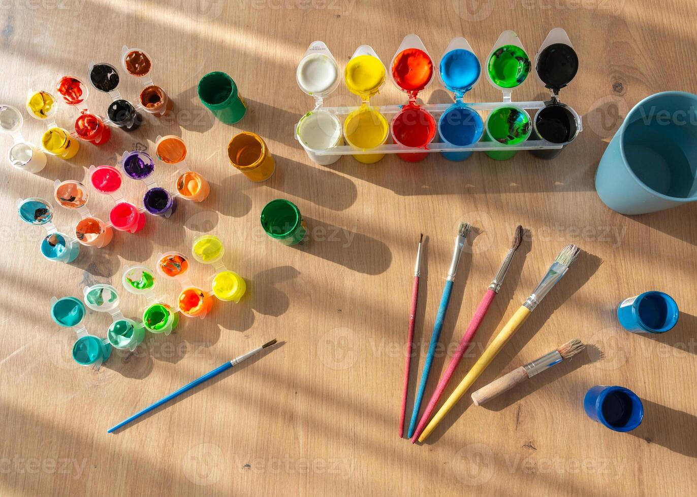 A set of finger paints, gouache, and brushes is arranged on a wooden table. Natural sunlight from the side casts soft shadows, emphasizing the artistic tools and vibrant colors photo