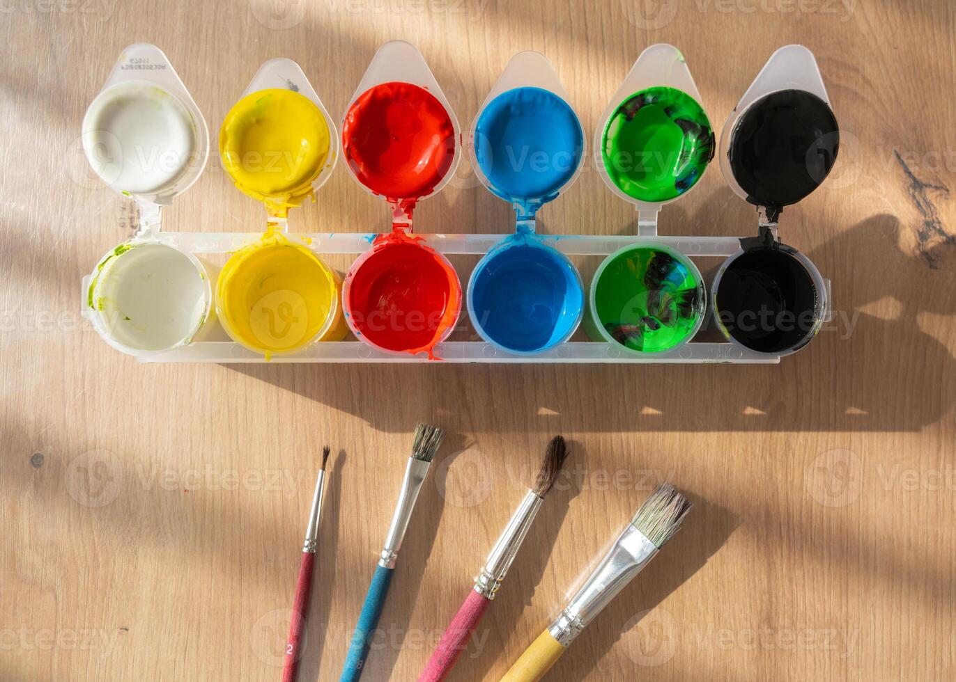Bright finger paints, gouache, and brushes are spread out on a wooden surface. Side sunlight enhances the vivid colors and details, making the scene feel warm and inspiring photo