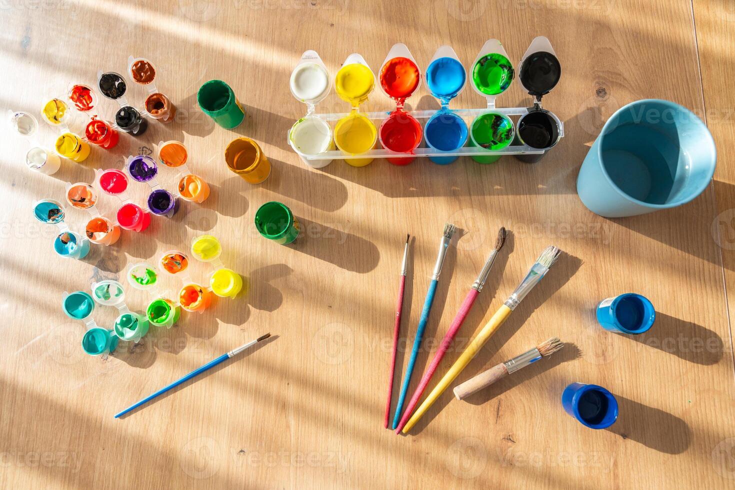 Finger paints and gouache with brushes are scattered on a wooden table. Sunlight from the side highlights the textures and colors, creating a warm and creative atmosphere photo