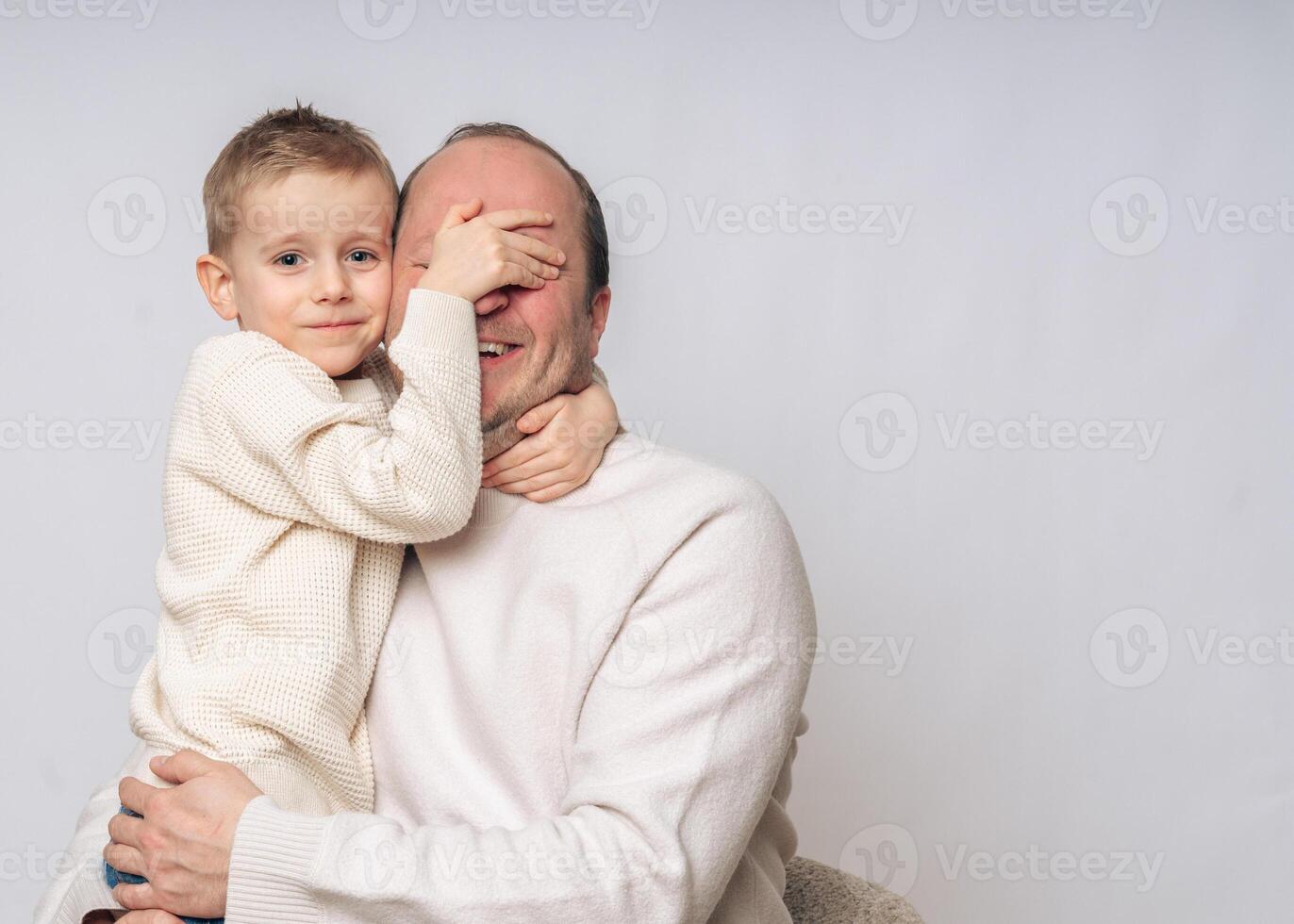 boy 4-5 years old boy sitting on daddy's lap boy covers daddy's eyes with his hand, the boy laughs, both of European appearance blond with blue eyes, studio shooting, dressed in light-colored clothes photo