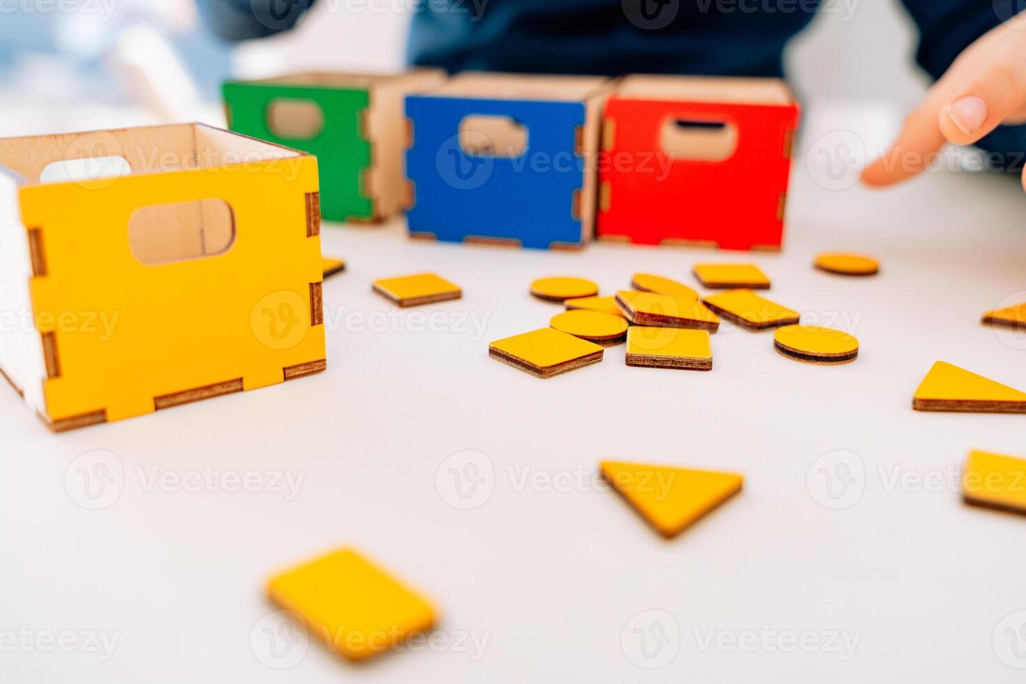 The little pupil arranges multicolored geometric shapes into the corresponding boxes. red, yellow, green and blue. yellow geometric shapes are scattered on the table. This Montessori-inspired photo