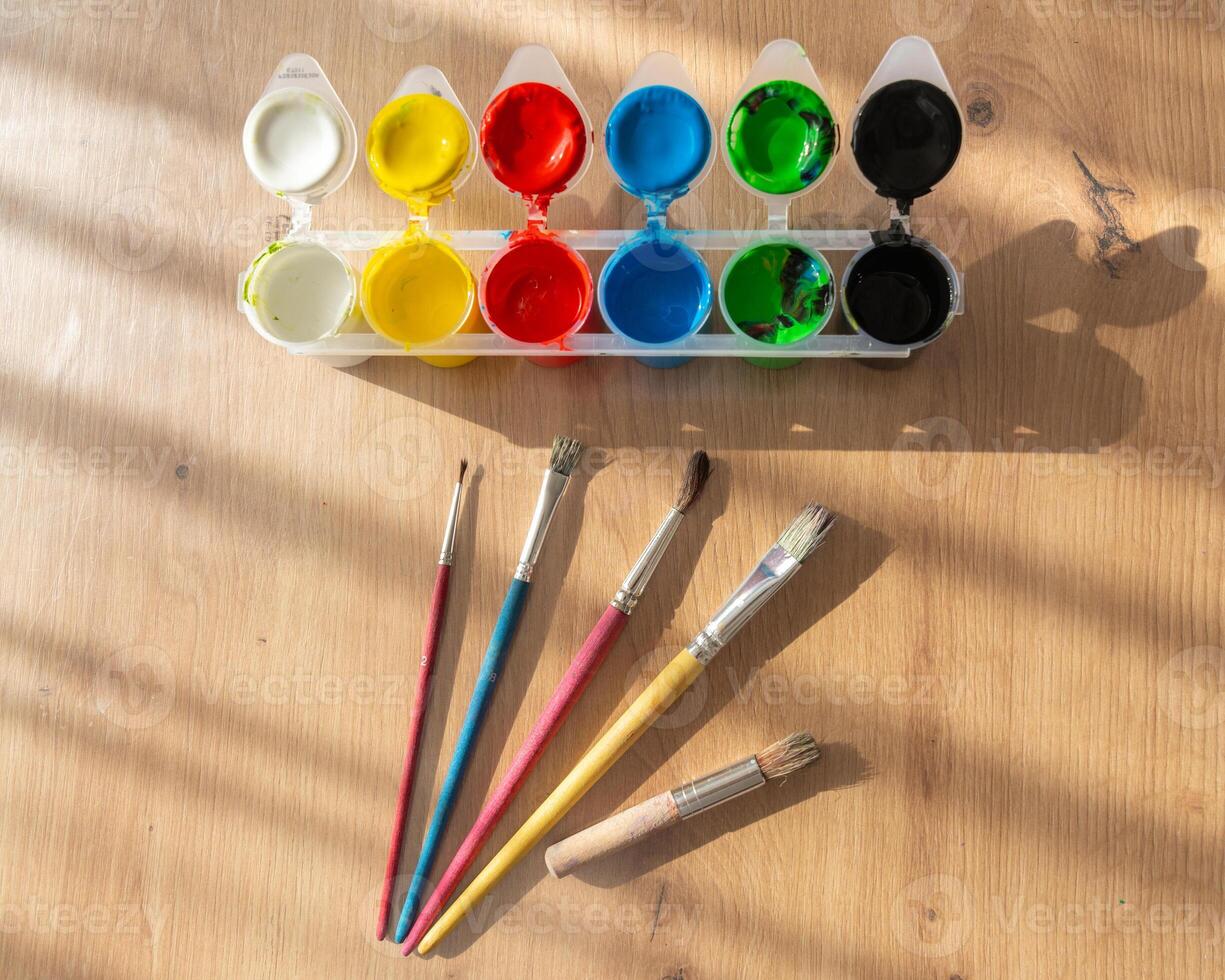 Finger paints or gouache and brushes spread out on a wooden table and side natural light from the sun photo