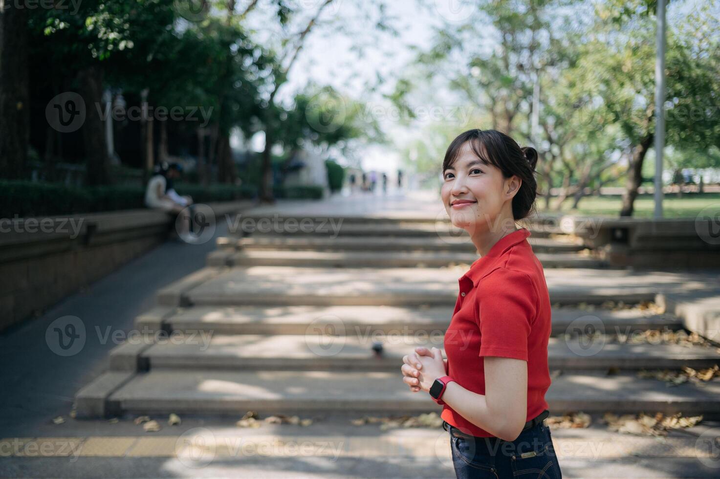 asiático alegre mujer en rojo camisa soportes al aire libre en soleado día, rodeado por arboles y pasos en parque me gusta ajuste foto