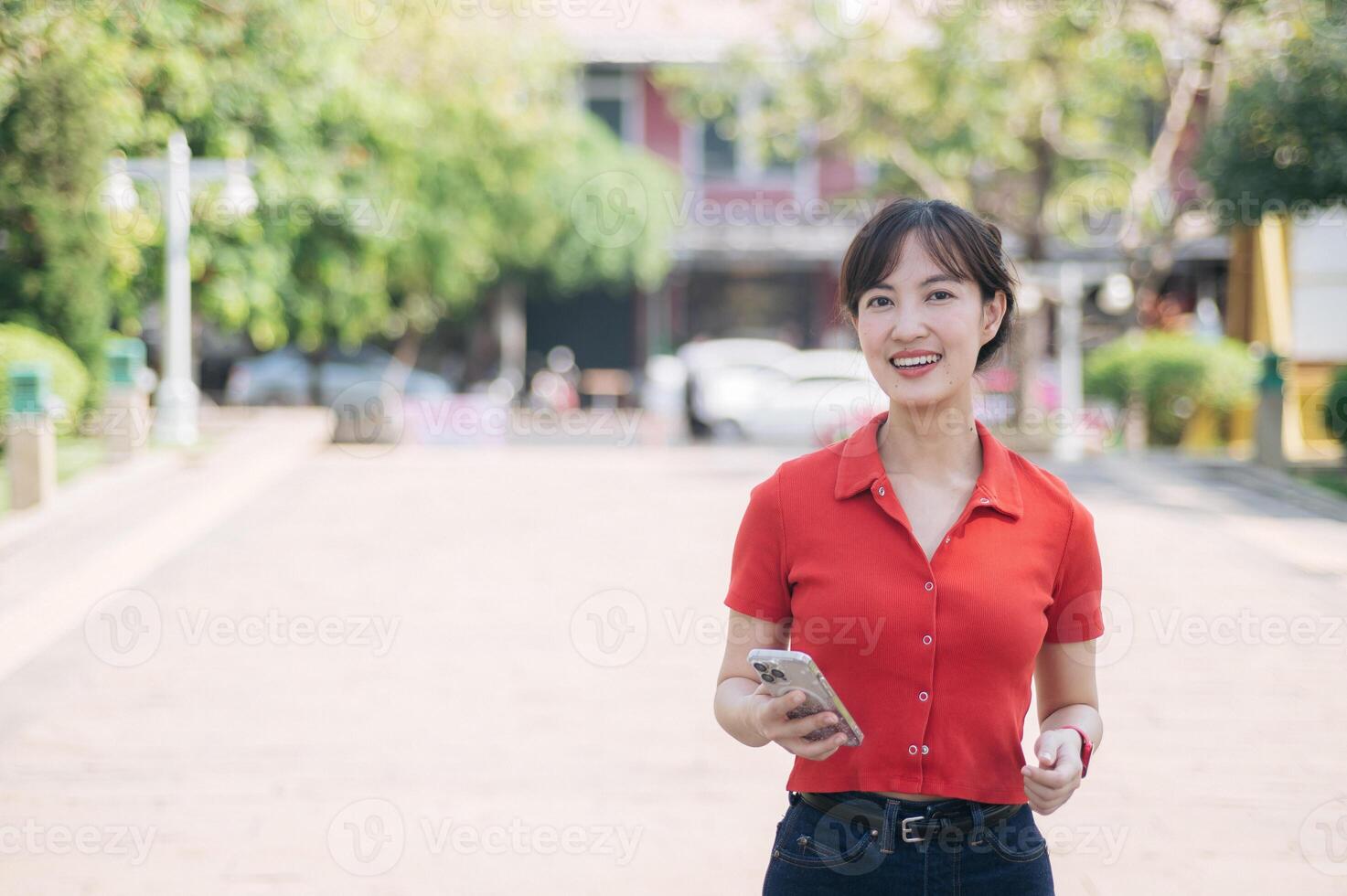Asian cheerful woman in red shirt holding smartphone and earbuds, standing outdoors on sunny day with blurred background of trees and buildings photo