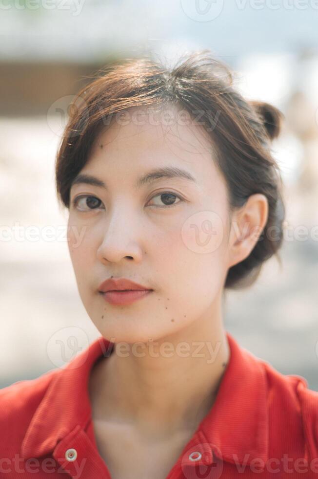 Asian thoughtful woman in red shirt gazes directly at camera, with soft natural light highlighting her features photo