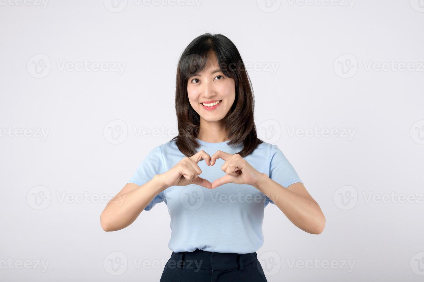 cheerful Asian woman in light blue shirt making heart shape with her hands against plain background photo