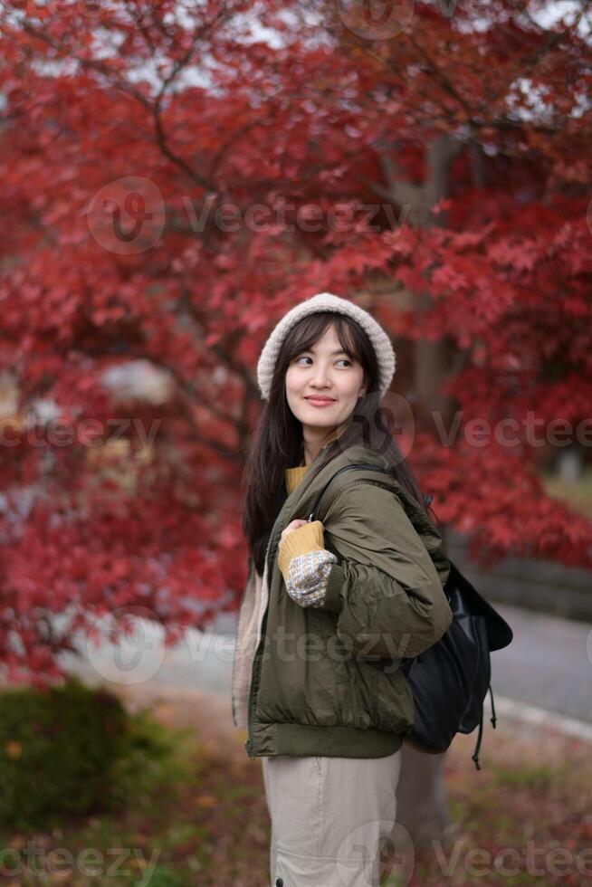 Asian woman in green jacket and hat standing in front of red maple tree photo