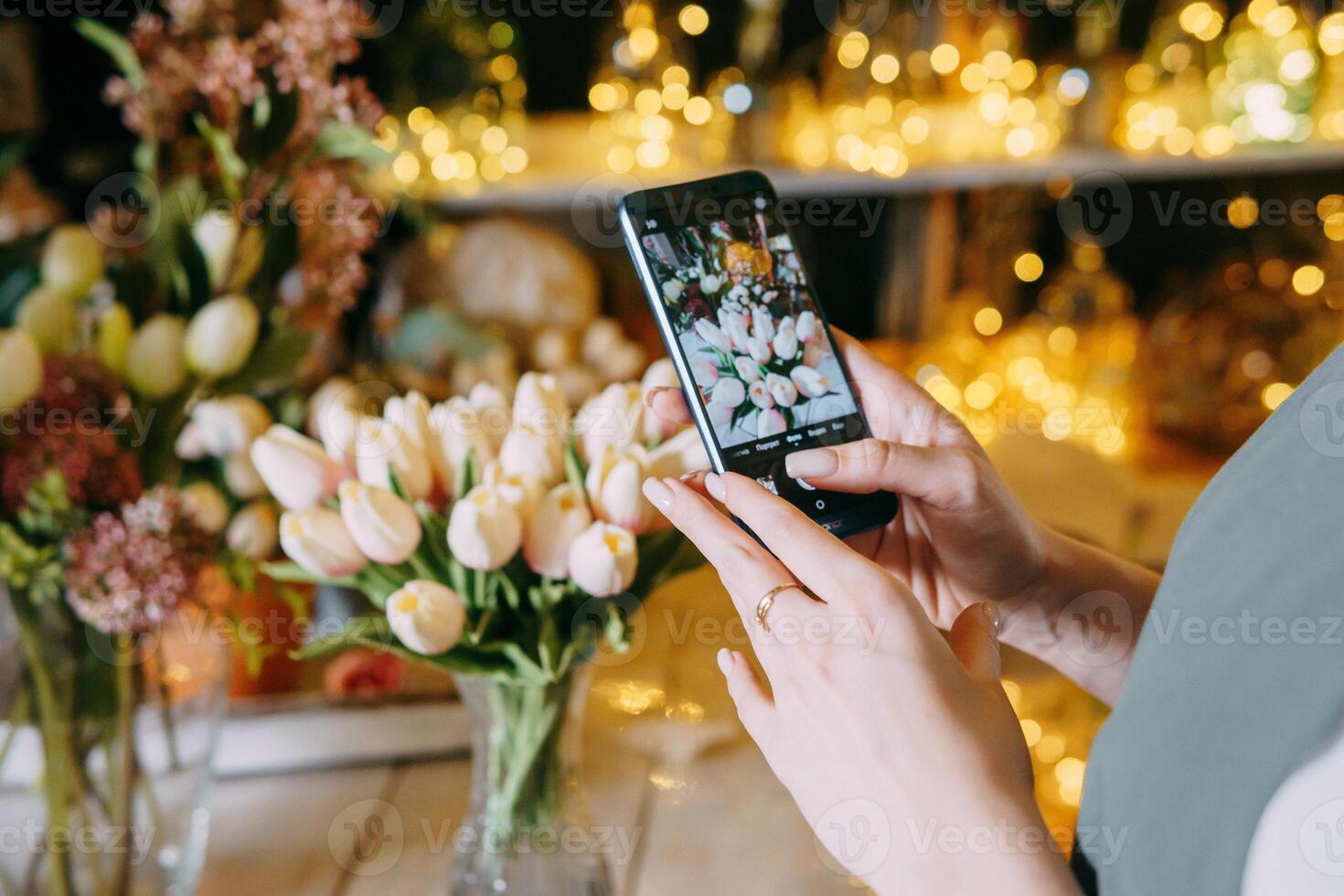 Close-up of person photographing bouquet of white tulips using smartphone in floral arrangement shop. Background features beautiful lighting and floral decor, creating warm. photo