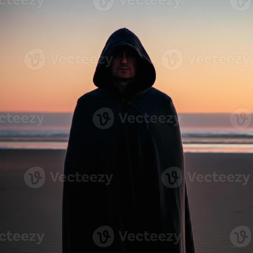 Mysterious hooded figure standing on a quiet beach at twilight, evoking secrecy, solitude, and an air of mystery photo