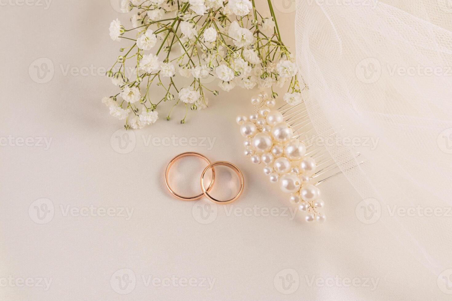 Two gold wedding rings, a pearl hairpin, a delicate champagne veil against a background of beige satin with a bouquet of white flowers. postcard photo
