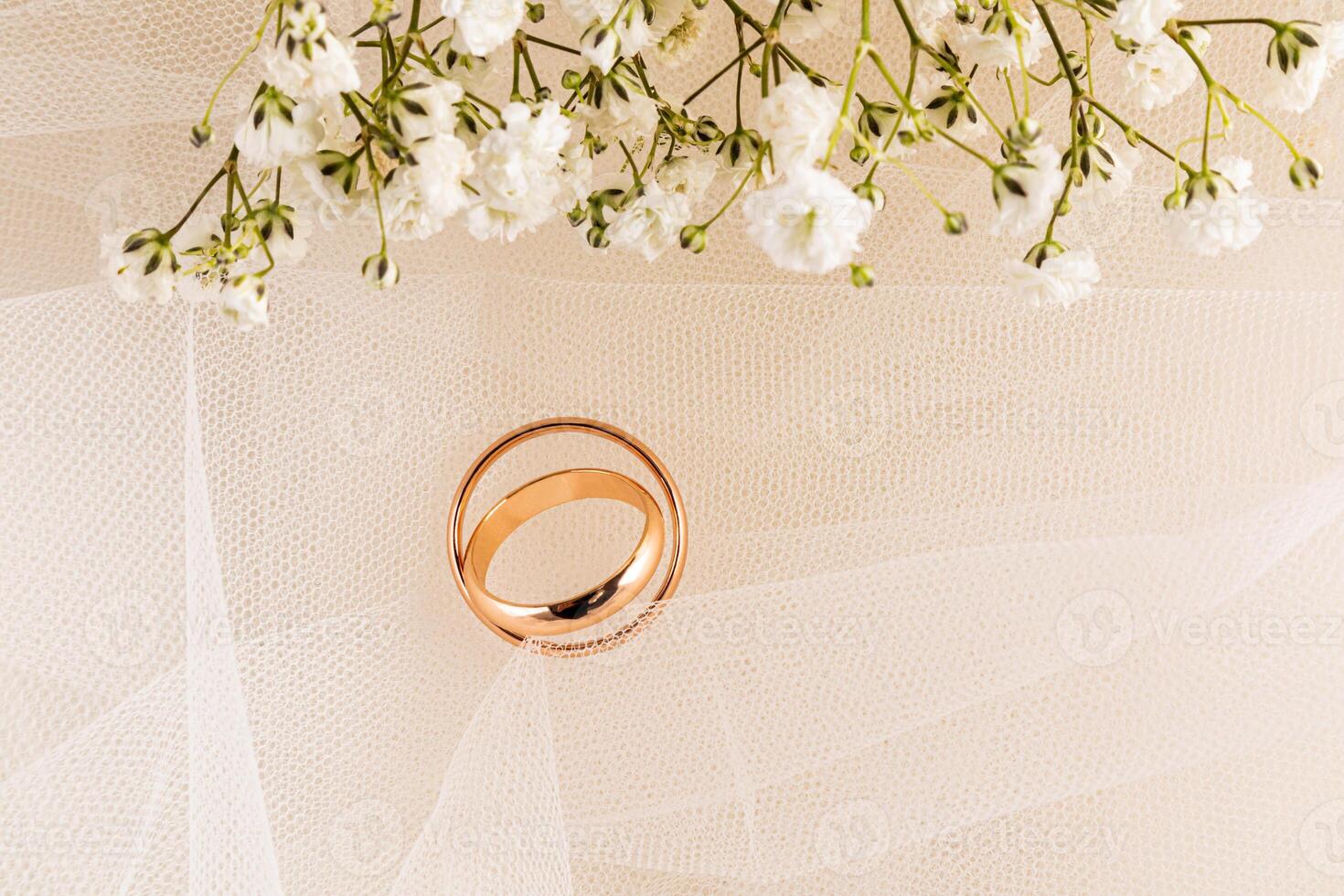 A pair of gold wedding rings delicately lying on the beige veil of the bride with a border of white gypsophila flowers. top view. A copy space photo