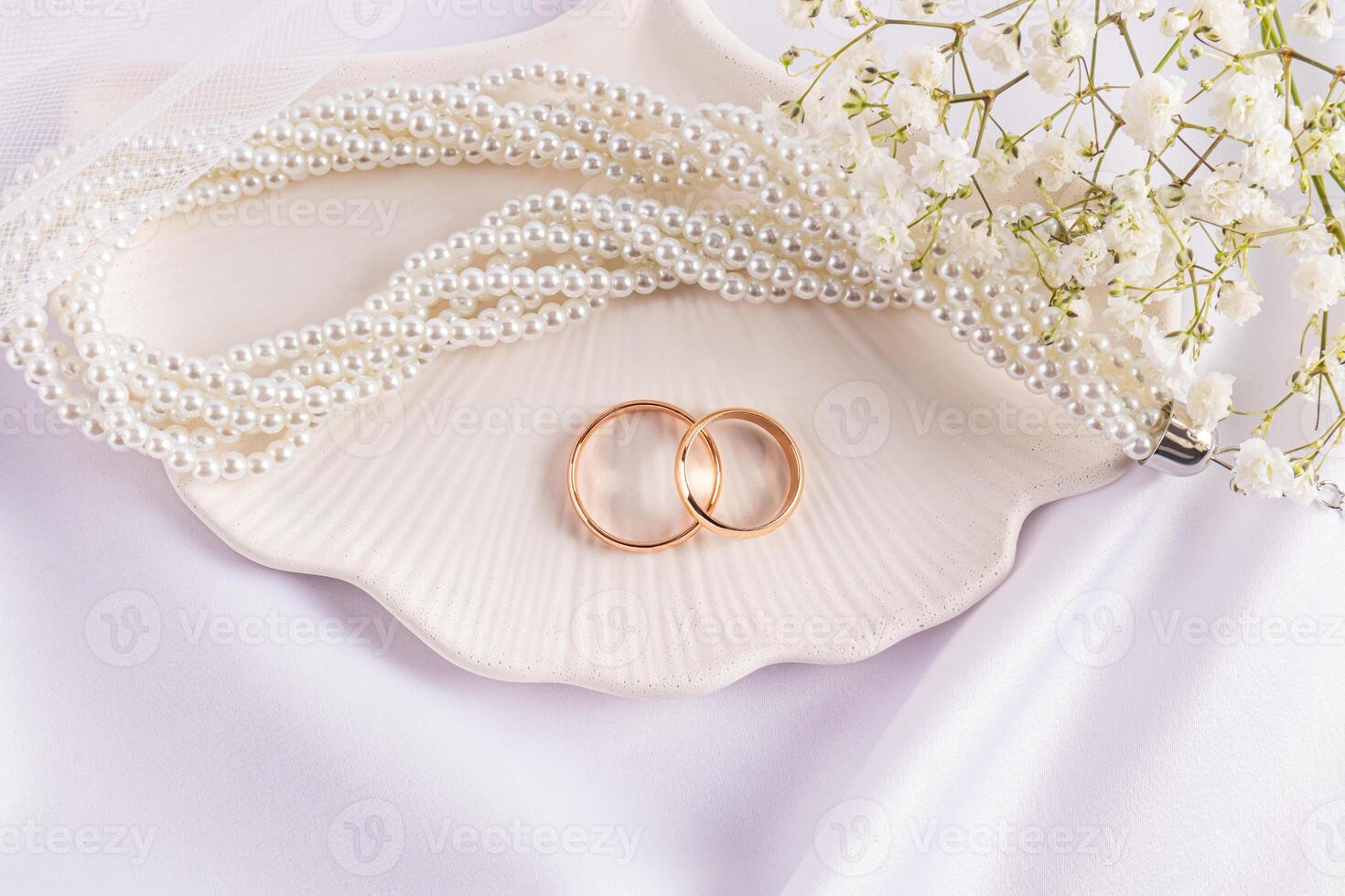 Two gold classic wedding rings are sitting on a decorative chic tray with pearl necklace and white gypsophila flowers. Delicate wedding card. layout. photo