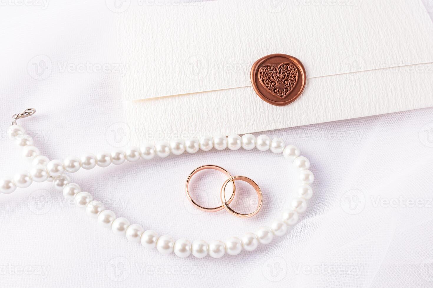 Wedding Arrangement with Wedding Rings, Pearl Beads, Envelope Invitation with Stamp on White Satin Background. top view. wedding layout, design. photo