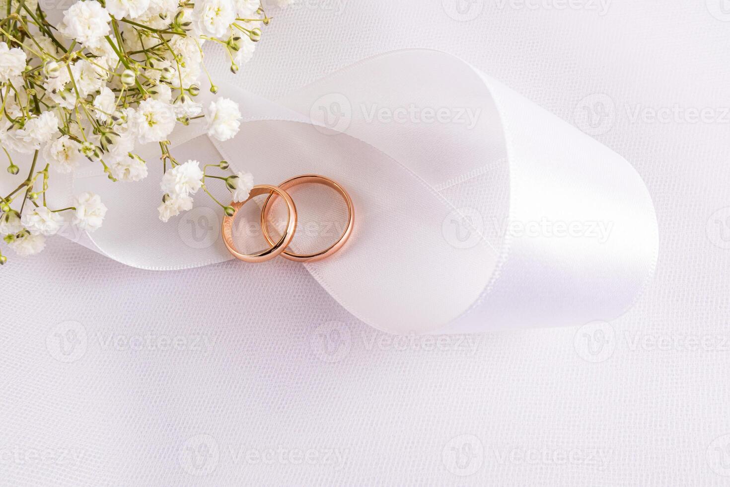 Two gold wedding rings lie on a chic satin white ribbon on white satin with delicate gypsophila flowers. top view. A copy space. postcard, invitation. photo